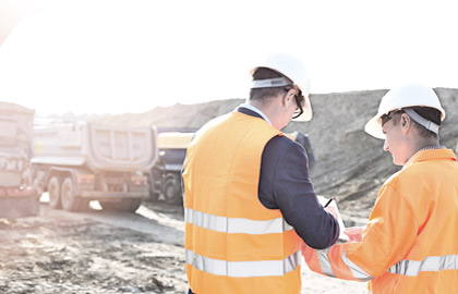 Bilde fra en utendørs arbeidsplass. To arbeidere med hvit hjelm og oransje vester står med ryggen til. I bakgrunnen skimtes tre lastebiler for transport av stein.